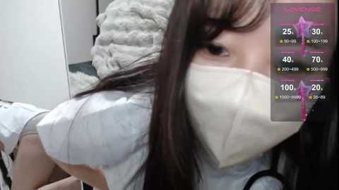 A close-up video of an Asian woman with long, dark hair, wearing a white face mask and a light blue shirt, lying on a bed with a white blanket.