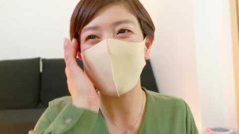 Media: Video of an Asian woman with short brown hair, wearing a green top and beige face mask, talking on a phone, against a blurred background of a black couch and white wall.