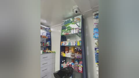 A video of a modern, well-lit pharmacy aisle with white walls, a CCTV camera, and shelves filled with various medications and health products.