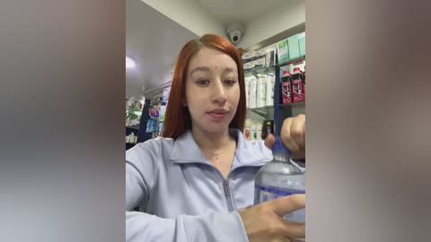 Video of a young woman with red hair, wearing a light blue zip-up jacket, holding a water bottle in a brightly lit pharmacy aisle.