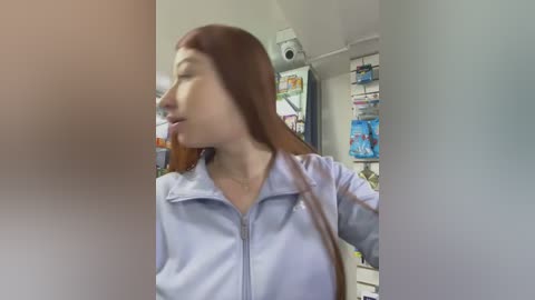 A video of a fair-skinned woman with long red hair, wearing a light blue lab coat, standing in a pharmacy aisle filled with shelves of medication boxes.