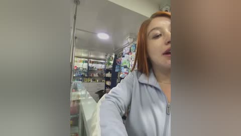 A video of a red-haired woman in a pharmacy, wearing a light blue jacket, taking a selfie. The background shows shelves stocked with medicine.