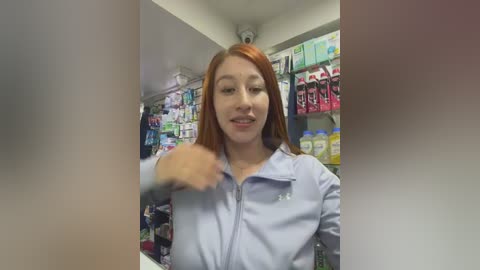 A video of a smiling Asian woman with long red hair, wearing a light blue shirt, standing in a brightly lit pharmacy with shelves filled with products.