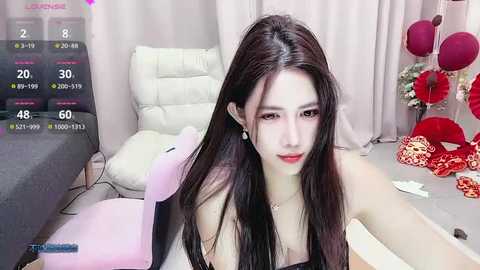 Media: A video of an East Asian woman with long, dark hair, fair skin, and red lipstick, sitting in a white chair, wearing a black dress, in a cluttered room with pink curtains, a TV, and red decorations.