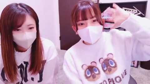 Media: Video of two East Asian women with long, straight brown hair wearing white masks and matching white sweaters with bear graphics, sitting indoors. One woman styles the other's hair.