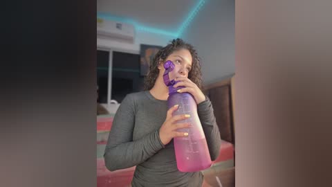 Video of a curly-haired woman with light brown skin, wearing a gray long-sleeve shirt, drinking from a pink water bottle, in a dimly lit bedroom with blue LED lights.