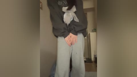 Media: A video of a person with light skin and long black hair, wearing a dark sweater, light scarf, and light blue pants, standing in a dimly lit, cluttered room with beige walls.