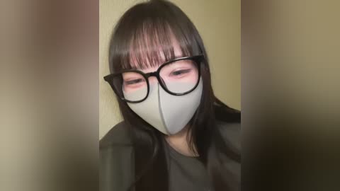 Media: Video of a person with long, dark hair, wearing black-rimmed glasses and a light gray face mask, set against a blurred background.