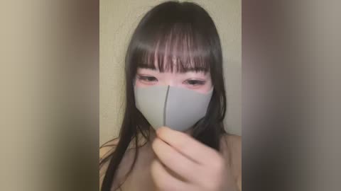 Media: Video of an Asian woman with long black hair and bangs, wearing a light gray face mask, covering her nose and mouth, holding the mask with her right hand. Background is a plain, light beige wall.