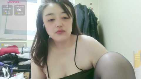 Media: Video of a plus-sized Asian woman with long black hair, wearing a black spaghetti-strap top and black stockings, sitting in a cluttered room with clothes and a TV screen displaying \"LIVE\" in the background.