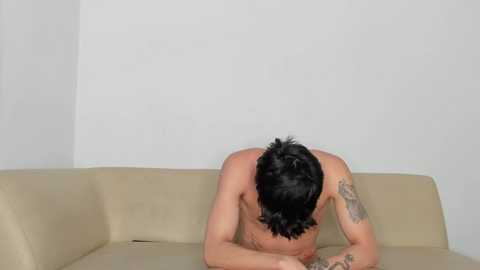 Media: Video of a shirtless man with short black hair, tattoos on his arms, lying face down on a beige couch against a plain white wall, appearing contemplative.