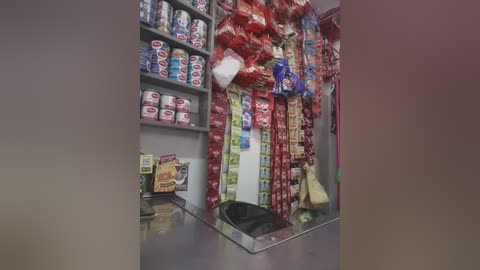 Video of a small convenience store with shelves filled with snacks and drinks, including numerous bags of chips and boxes of cookies, a cash register, and a black trash bin.