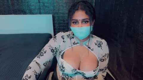 Media: A video of a South Asian woman with curly hair, wearing a turquoise face mask, a floral-patterned top that exposes cleavage, and a black bra. She's seated on a bed in a dimly lit room with dark, textured walls.