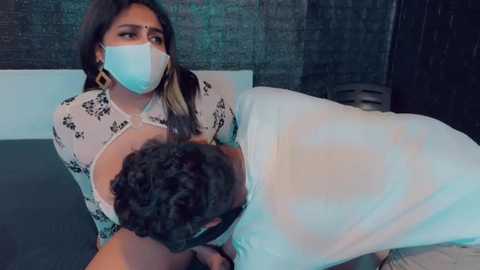 Media: Video of a woman in a mask and floral-patterned top cradling a man with curly hair in a hospital setting, with a dark brick wall and blue bedsheet.