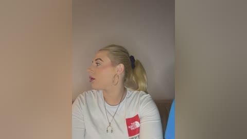 Media: Video of a fair-skinned, blonde woman with a ponytail, wearing a white T-shirt with a red patch, sitting in a beige room.