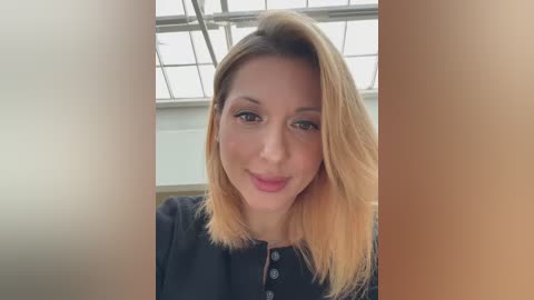 Media: Video of a smiling Caucasian woman with shoulder-length, light brown hair, wearing a black top, against a bright, modern office backdrop with large windows.