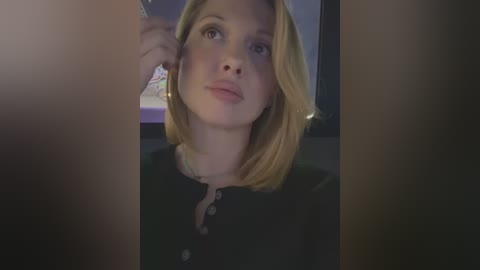 Media: Video of a young woman with shoulder-length blonde hair, light skin, and a neutral expression, wearing a black top, taken indoors with blurred background.