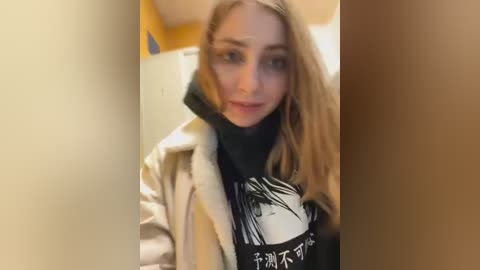 Media: A video of a young woman with long blonde hair, fair skin, and blue eyes, wearing a black turtleneck and beige jacket, taken in a dimly lit hallway with yellow walls.