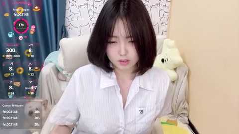 A young Asian woman with straight black hair, wearing a white striped blouse, sits at a desk in a cozy room with pastel decorations and a plush toy.