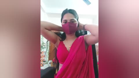 Media: A video of a woman with medium skin tone, wearing a bright red sari, a red face mask, and a gold necklace, posing with her arms raised, indoors with a white ceiling fan visible.