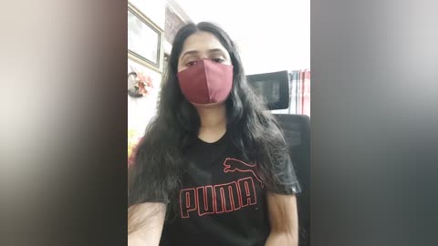 Media: A video of a woman with long, dark hair, wearing a red face mask and black PUMA T-shirt, seated indoors, blurred background with framed pictures and a chair.