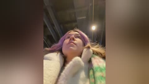 Media: Video of a young woman with light skin and pastel pink hair, wearing a green and white checkered coat, gazing upwards in an indoor setting with a dimly lit ceiling and visible industrial pipes.