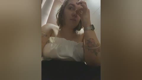 Media: A video of a woman with light skin and shoulder-length brown hair, wearing a white strapless dress, leaning against a white curtain in a dimly lit room. She has tattoos on her arm and is holding her head with a watch on her wrist.