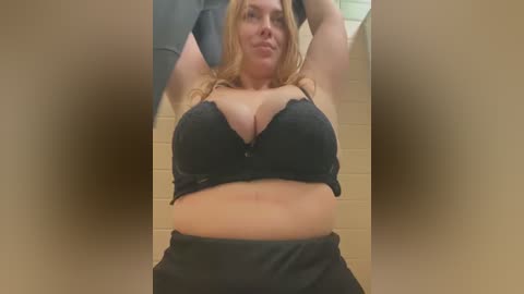 Media: Video of a plus-sized woman with fair skin and long blonde hair, wearing a black lace bra that accentuates her large breasts, and black pants. She has a confident smile, standing in a tiled bathroom with beige walls.