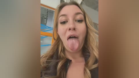 Media: Video of a young Caucasian woman with long blonde hair, wearing a dark jacket, sticking out her tongue, taken indoors with a blurred background.