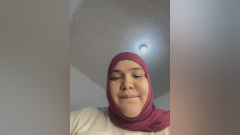 Video of a smiling woman with medium brown skin and a maroon hijab, standing in a room with a textured ceiling and a small light fixture.