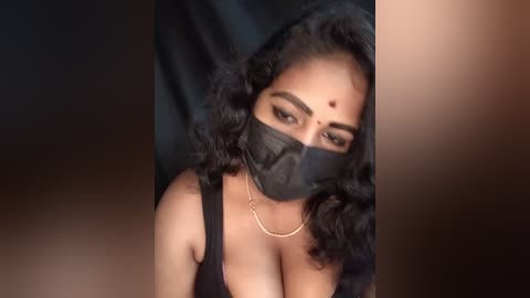 Media: Video of a South Asian woman with medium-dark skin, wearing a black face mask, black sleeveless top, and a thin gold necklace, with long black wavy hair and a red bindi on her forehead, set against a dark background.