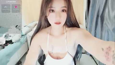 Media: A video of a young Asian woman with long brown hair, wearing a white halter top, sitting on a bed with messy clothes. She has a tattoo on her left arm.