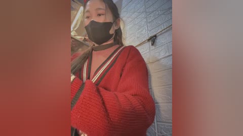 Media: Video of an Asian woman with long black hair, wearing a red knit cardigan, black face mask, and black choker. She stands against a white tiled wall, indoors.