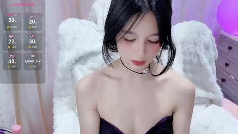 Video of an East Asian woman with fair skin and black hair, wearing a black strapless top, sitting on a white bed in a softly lit room.