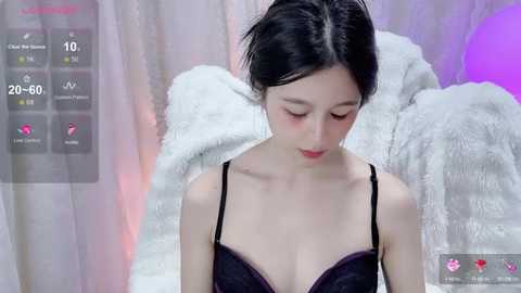 A video of an Asian woman with fair skin and black hair, wearing a black lace bra, standing against a white and purple background. She appears serene, with a soft expression.