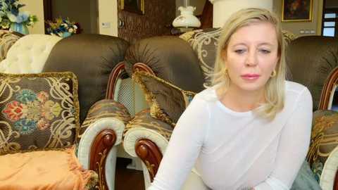 Video of a blonde woman with fair skin in a white top, sitting on a plush, ornate sofa in a cozy living room, surrounded by patterned cushions.