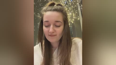 Video of a young woman with long brown hair, wearing a white top, and a delicate necklace, gazing downward in a serene indoor setting with string lights in the background.