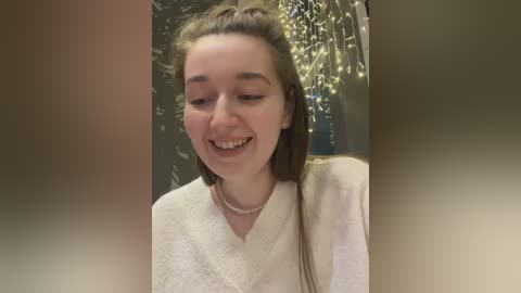 A video of a young, smiling Caucasian woman with light skin and long, straight brown hair. She wears a cream-colored sweater and a simple pearl necklace. The background features a blurred, decorative Christmas tree with fairy lights.