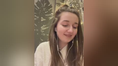 Video of a young woman with long, straight brown hair, wearing headphones, looking down with a soft smile, against a background of a Christmas tree and a warm, cozy room.