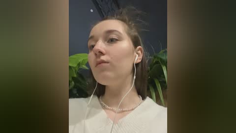 A video of a young woman with light skin and brown hair, wearing headphones, a white blouse, and a pearl necklace, in a room with green plants in the background.