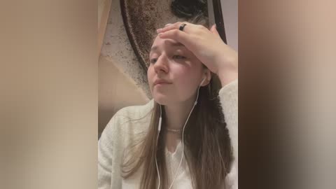 Video of a young woman with long, straight brown hair, wearing a white shirt and gray cardigan, listening to earphones with one hand on her forehead, in a beige bathroom with a granite countertop.