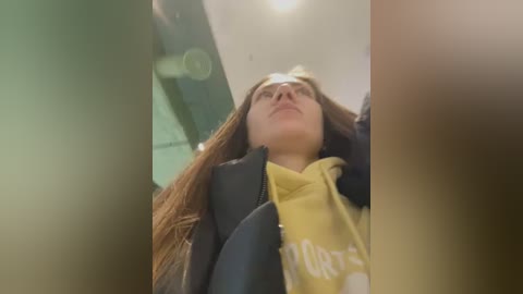 A video of a young woman with long brown hair, wearing a yellow hoodie and black jacket, looking upward with a contemplative expression. The background is blurred, with hints of a glass door and ceiling lights.