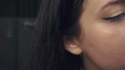 A close-up video of a young woman with fair skin, dark hair, and long eyelashes, partially in focus. Her profile is angled towards the left, revealing her ear. The background is blurred, suggesting an indoor setting.