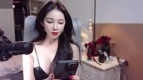 Media: Video of an Asian woman with long black hair, wearing a black leather top, recording herself on a smartphone, in a modern bedroom with a white dresser and red flowers.