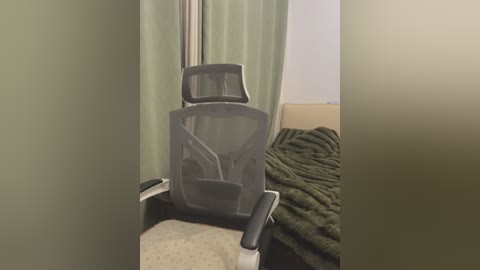 Video of a modern, gray mesh office chair with adjustable armrests, placed in a small, dimly lit room with beige walls, a green curtain, and a dark green blanket draped over a bed.