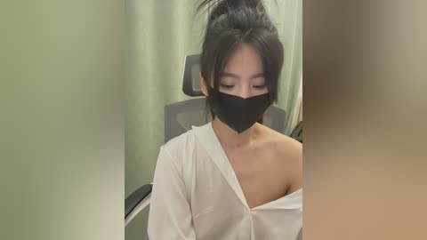 Media: Video of an East Asian woman with medium skin tone and black hair in a bun, wearing a black mask and white off-shoulder shirt, seated in a dimly-lit room with green walls and a gray chair.