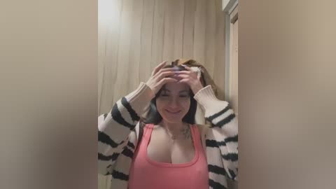 Video of a young woman with fair skin and long brown hair, smiling, adjusting her hair in a bathroom with beige walls and a white door. She wears a pink tank top and a striped cardigan.