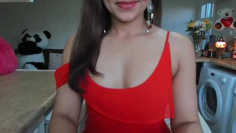 Media: Video of a young woman with long dark hair in a red off-shoulder dress, standing in a cozy kitchen with a panda plush, a white washing machine, and flowers in the background.