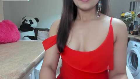 Media: A video of an Asian woman with long black hair, wearing a red off-shoulder dress, standing in a modern kitchen with a panda plush toy, a washing machine, and pink fluffy cushions.