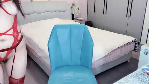 Video of a modern bedroom featuring a light grey upholstered bed with white bedding, a blue chair, and a red lingerie-clad woman standing beside it.